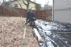 Lay out the deck area with stakes and string, and lay down plastic sheeting to prevent weeds and unwanted vegetation from growing beneath the deck.
