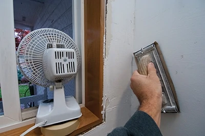 A hand sander was used to smooth out bumps and ridges in the finish. The sander holds precut sand paper. Notice the small fan positioned in the window to exhaust dust from the room.